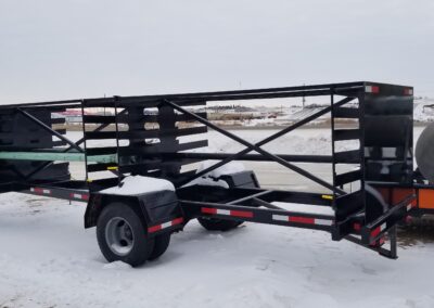 Heavy-duty black steel utility trailer with cross-braced sides, parked on snowy lot, ideal for pipe or equipment hauling.