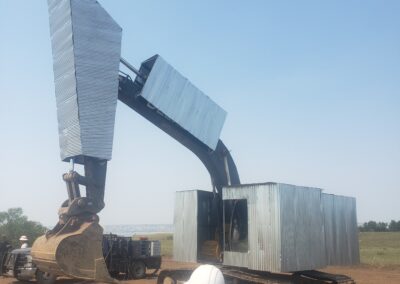 Modified excavator with corrugated metal shielding at construction site, workers in hard hats inspecting heavy equipment.