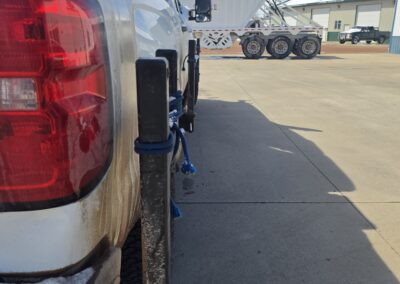 White 4x4 pickup with tow hitch posts and blue rope on snowy bumper in industrial yard; dump trailer in background.
