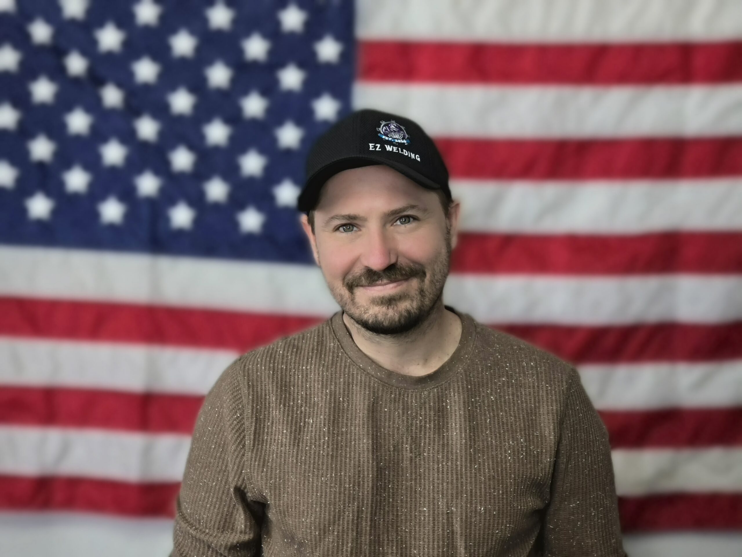 Smiling man in brown sweater wearing EZ Welding cap before American flag backdrop