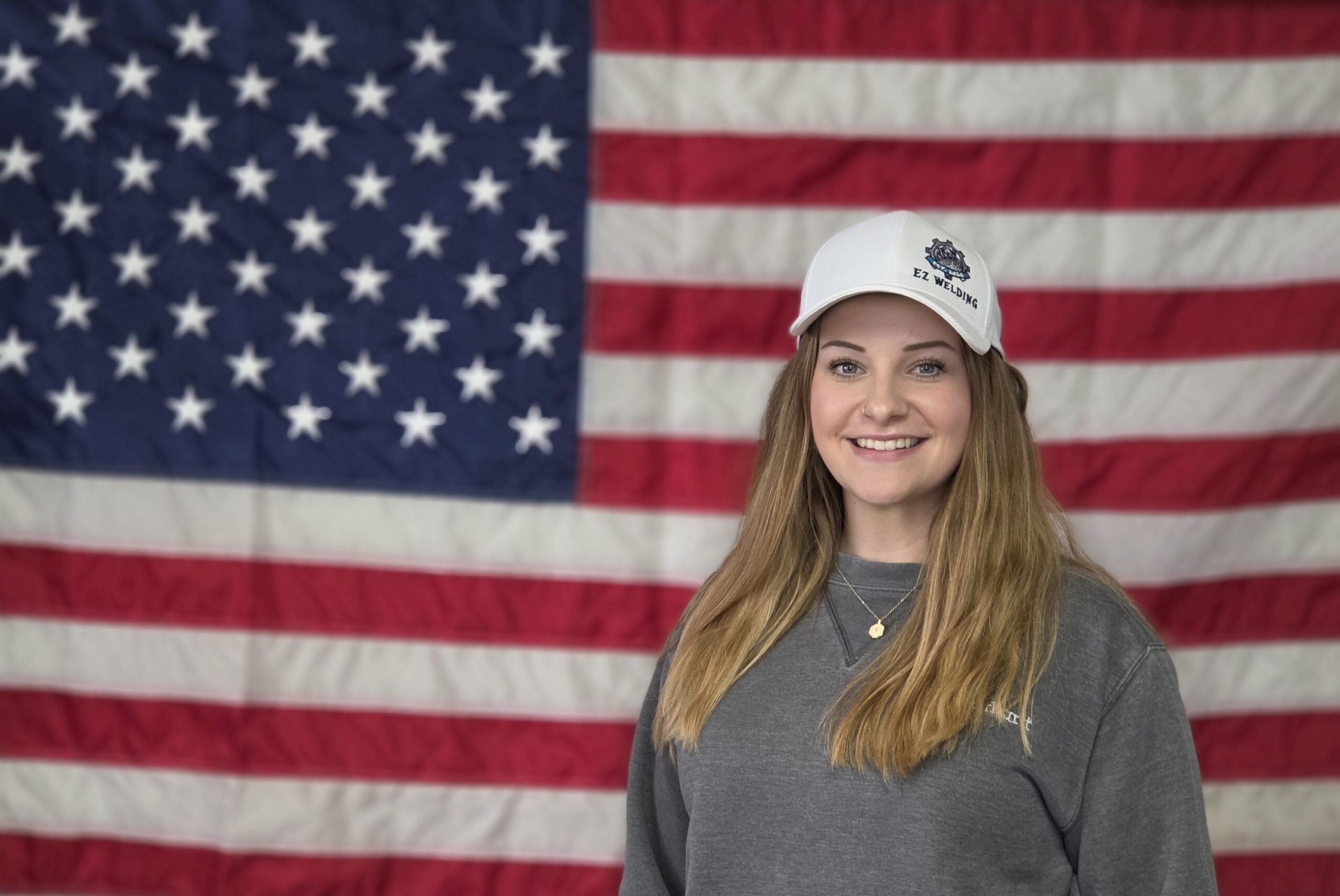 Smiling woman in EZ Welding hat stands before American flag, patriotic USA background.