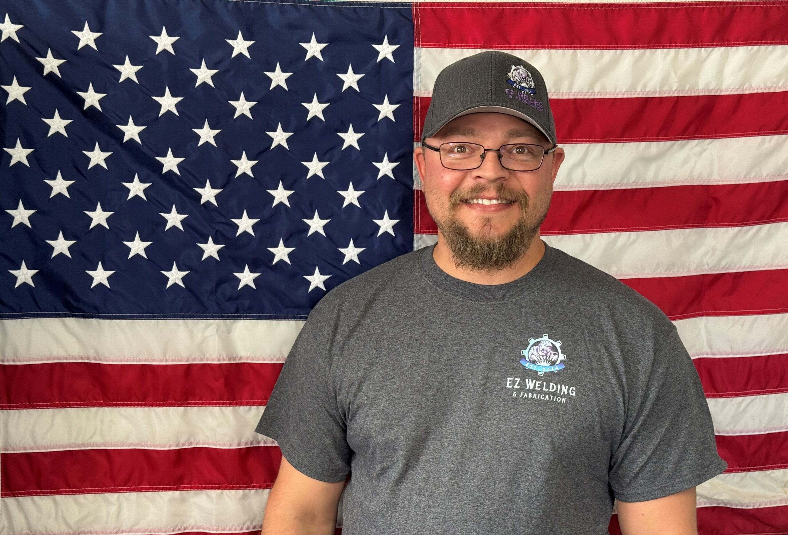 Smiling welder wearing EZ Welding & Fabrication shirt and cap in front of the American flag